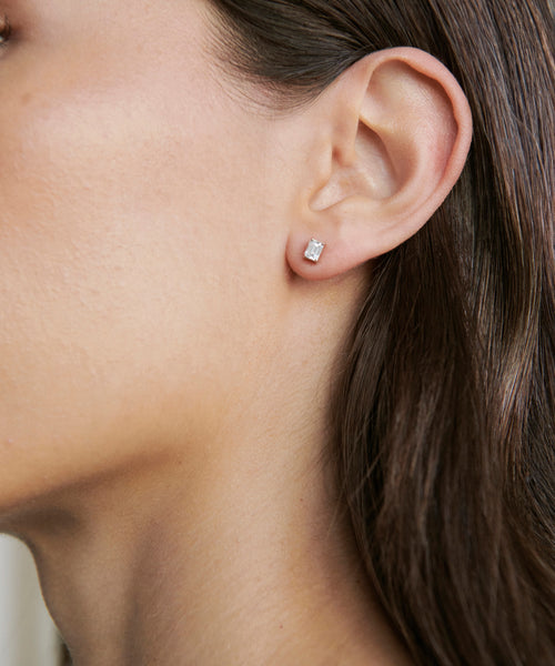 Close-up of a womans ear adorned with Ivy Emerald Cut Studs in white gold. The neutral background emphasizes the earrings elegant rectangular shape, while her brown hair is tucked neatly behind her ear.