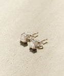 The Ivy Emerald Cut Studs feature minimalist silver settings with rectangular clear gemstones, showcased on a textured beige surface.