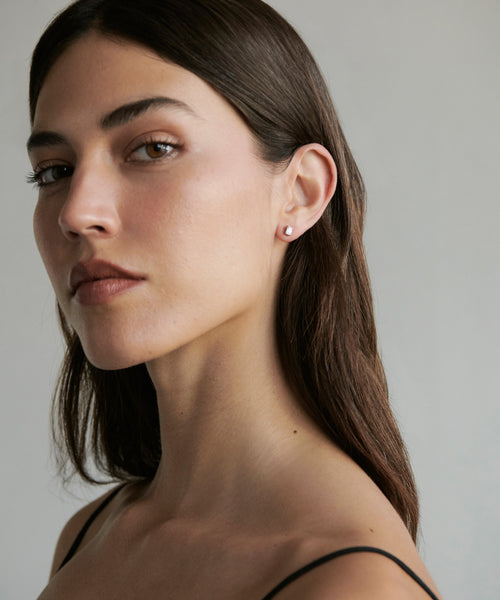 A woman with long brown hair and a black strap gazes calmly at the camera, wearing Ivy Emerald Cut Studs crafted from conflict-free diamonds and white gold, posed against a plain, light background.