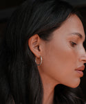 A woman with long dark hair and smooth skin wears Medium Eloise Hoops, shown in side profile as she looks down against a dark background.