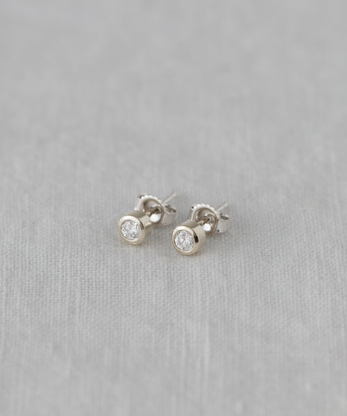The Stevie Diamond Studs feature minimalist round, clear diamonds set in white gold, shown on a light gray fabric background—a timeless design inspired by Jenni Kayne jewelry.