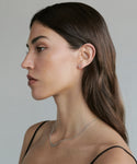 A woman with straight brown hair is shown in profile against a plain background. She wears small Stevie Diamond Studs earrings and a thin necklace, with minimal makeup and a calm, neutral expression.