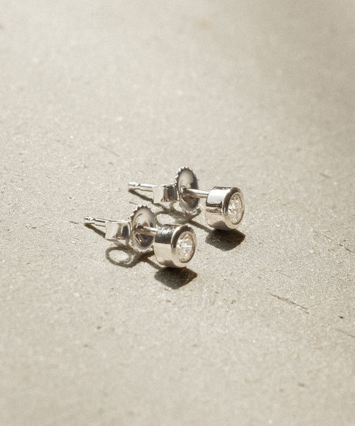 The Stevie Diamond Studs—small, round white gold earrings with clear gemstones—rest on a textured light surface in soft natural light, capturing the subtle elegance of Jenni Kayne jewelry.