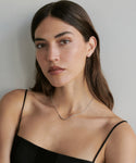 A woman with long brown hair and light skin wears a black spaghetti strap top, a delicate Jenni Kayne silver necklace, and Stevie Diamond Studs, looking calmly at the camera against a plain light background.