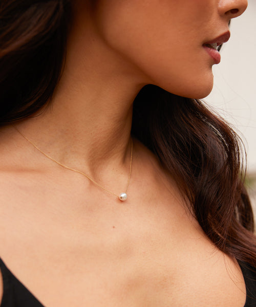 Close-up of a woman’s neck and lower face wearing the Philo Pearl Necklace, a 14k gold chain with a single pearl pendant. She has long dark hair and is dressed in a black top.