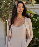 Wearing a light beige dress and cardigan, a woman with long dark hair stands outdoors by greenery and a white fence, her look completed by the Philo Pearl Necklace, which adds delicate elegance as she gazes calmly to the side.