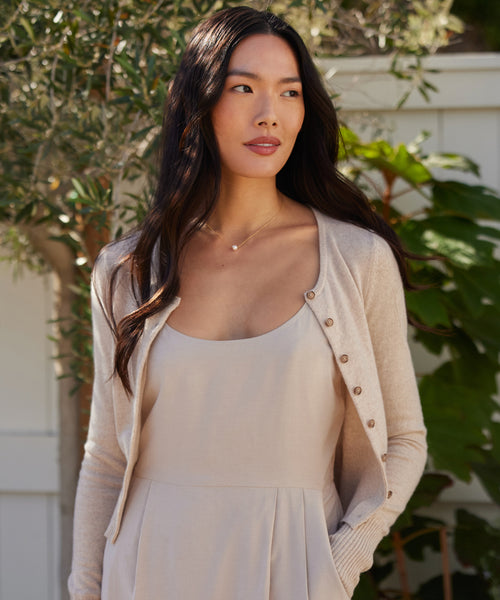 Wearing a light beige dress and cardigan, a woman with long dark hair stands outdoors by greenery and a white fence, her look completed by the Philo Pearl Necklace, which adds delicate elegance as she gazes calmly to the side.