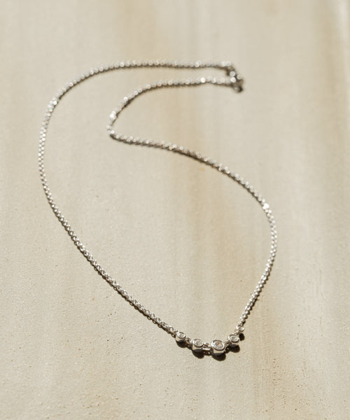 The Violette Diamond Necklace features a silver chain with five small round diamonds evenly spaced at the center, displayed on light beige textured fabric in natural light—a timeless, heirloom-inspired piece.