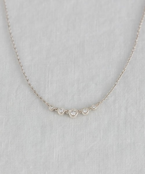 The Violette Diamond Necklace in white gold features a pendant with five small round diamonds in a row, displayed on a light gray textured fabric background.