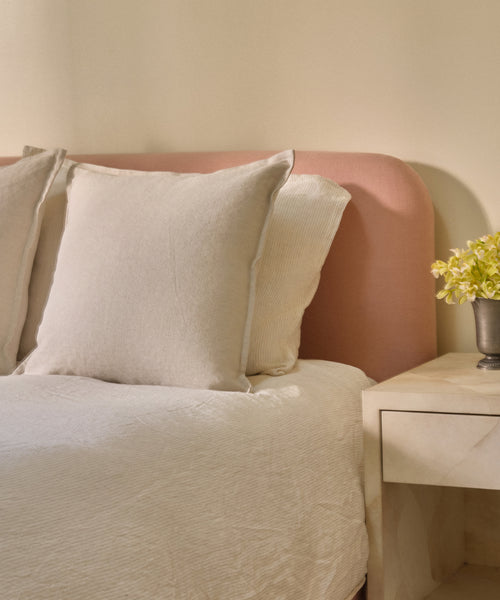 A neatly made bed with cream-colored bedding and an Adler Pillow in Belgian linen, a soft pink headboard, and a beige nightstand holding a small vase of yellow flowers against a light-colored wall.