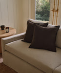 A beige sofa with two dark brown Adler Pillows is placed beside a round wooden side table holding a lit candle and a mug, positioned near a large window overlooking greenery.