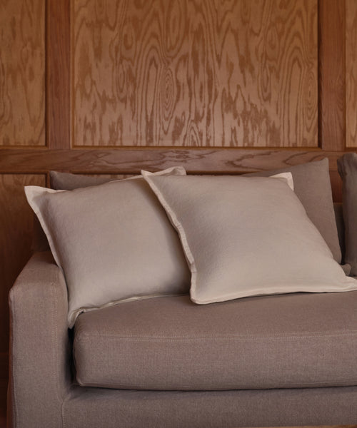A beige sofa with two light Adler Pillows is placed in front of a wooden-paneled wall showcasing natural wood grain.