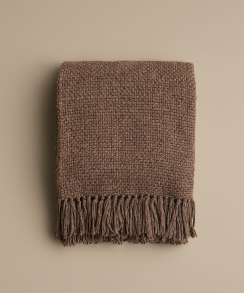 The Alpaca Basketweave Throw, featuring a folded, textured brown design with long fringes at the ends, is displayed on a plain beige background.
