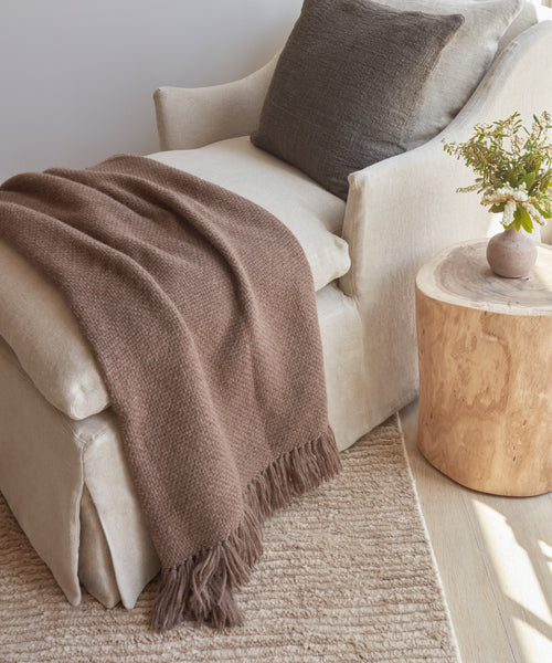 A cozy beige armchair with a brown Alpaca Basketweave Throw made in Peru and a dark cushion sits by a round wooden side table with flowers, atop a light woven rug in a softly lit room.