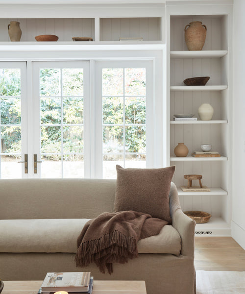 A cozy living room features a beige sofa, a brown pillow, the Alpaca Basketweave Throw made in Peru, a wooden coffee table with books, built-in pottery shelves, and large glass doors opening to a garden.