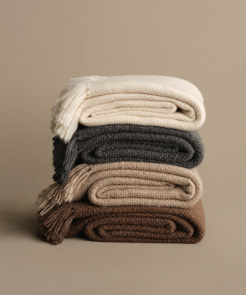 A stack of four folded, knitted blankets—including the Alpaca Basketweave Throw—in cream, dark gray, light beige, and brown rests on a neutral beige surface with a matching background.