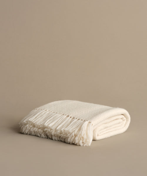 The Alpaca Basketweave Throw, handwoven in Peru from superfine alpaca, features fringed edges and an off-white hue. Elegantly folded on a beige surface, it brings subtle luxury and warmth to any space.