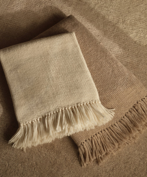 Two Alpaca Basketweave Throws, handwoven in Peru from superfine alpaca—one cream, one light brown with fringed edges—are neatly folded and overlapping on a soft, neutral-toned surface.