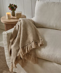 The Alpaca Basketweave Throw, a beige handwoven Peruvian alpaca blanket with fringe, is draped over a light sofa. Nearby, a wooden side table with a lit candle and small vase adds to the cozy scene.