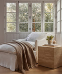 A cozy white armchair with an Alpaca Basketweave Throw and cushion sits by large windows; nearby, a travertine side table holds a vase of greenery and a lit candle. Sunlight streams through the trees outside.