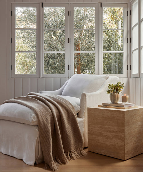 A cozy white armchair with an Alpaca Basketweave Throw and cushion sits by large windows; nearby, a travertine side table holds a vase of greenery and a lit candle. Sunlight streams through the trees outside.