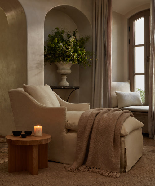 A cozy neutral-toned living corner features a cream armchair draped with the Alpaca Basketweave Throw, a round wooden side table with candle and cups, an arched alcove with greenery, and a window bench framed by soft drapes.