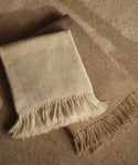 Two Alpaca Basketweave Throws—one cream and one light brown—are neatly folded and overlapping on a textured beige surface. Handwoven in Peru from superfine alpaca, they offer exceptional softness and warmth.