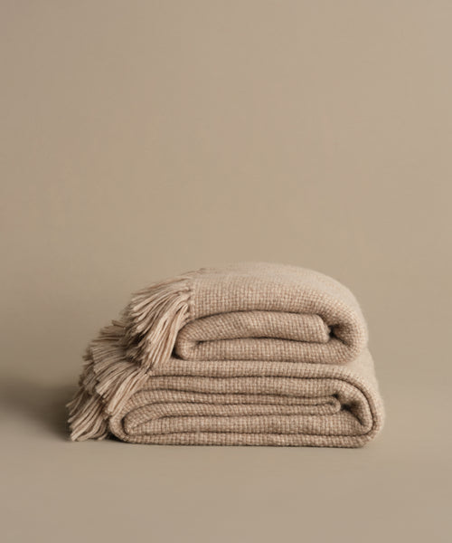 The Alpaca Basketweave Throw, handwoven in Peru from superfine beige alpaca wool with fringed edges, is folded on a matching surface against a plain beige background.