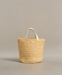 The Capri Bag is a handcrafted woven straw tote with two short white handles, shown against a plain neutral background.