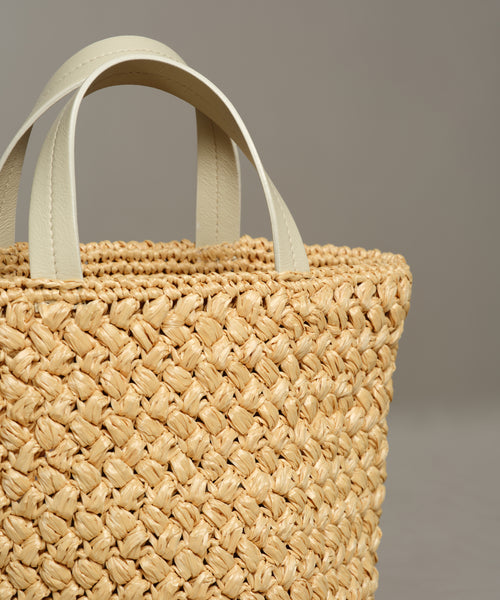 Close-up of the Capri Bag, showcasing its woven straw body and two short, white leather handles against a plain, light gray background.