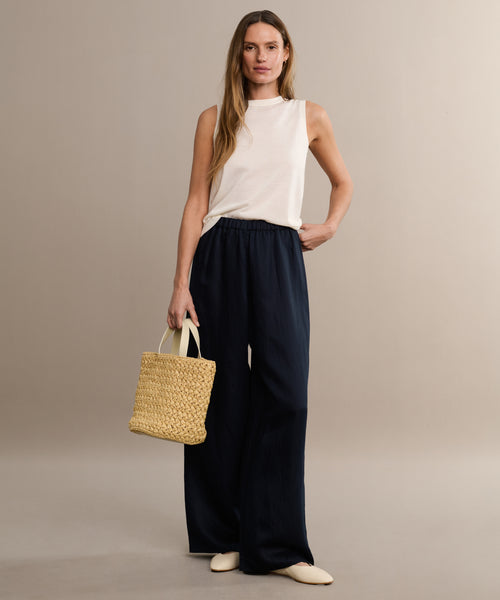 A woman with long, wavy hair stands against a neutral background wearing a sleeveless white top, loose dark navy pants, and cream shoes. She holds the Capri Bag, a handcrafted woven straw accessory.