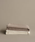 Two neatly folded blankets, including the luxurious Chateau Alpaca Coverlet in ivory and a beige throw, are stacked atop each other against a plain light brown background.