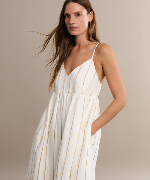 A woman with long brown hair wears the Cove Dress, a sleeveless casual dress with slender straps and thin gold stripes, standing with one hand in her pocket against a beige background.