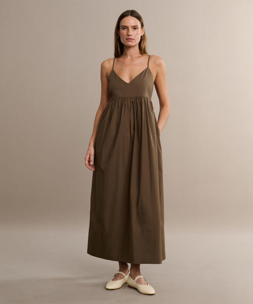 A woman stands against a plain beige background, wearing the Cove Dress—a sleeveless brown cotton design with thin straps and a gathered waist for a relaxed fit—paired with light-colored flat shoes.