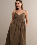 A woman stands against a neutral background wearing the Cove Dress—a sleeveless, brown, V-neck cotton dress with thin straps and pockets—looking slightly to the side for a touch of casual flair.
