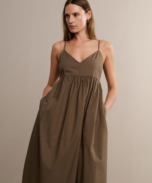 A woman stands against a neutral background wearing the Cove Dress—a sleeveless, brown, V-neck cotton dress with thin straps and pockets—looking slightly to the side for a touch of casual flair.