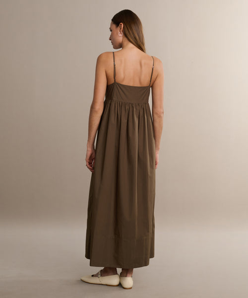 A woman with straight brown hair stands facing away, wearing the Cove Dress—a relaxed fit, brown cotton dress with thin straps—and cream-colored flat shoes against a plain beige background.