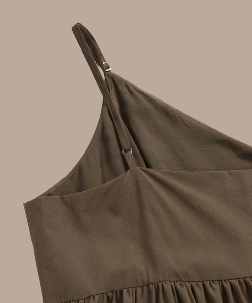 A close-up of the Cove Dress, a brown cotton dress with a relaxed fit, adjustable shoulder straps, and gathered waist detail, laid flat on a beige surface.