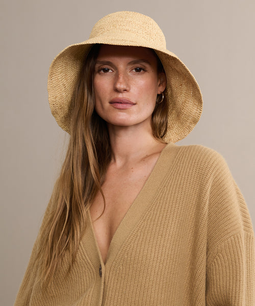 A woman with long brown hair wears the Crochet Raffia Sun Hat and a beige knit cardigan, gazing confidently at the camera against a plain light brown background.