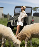 Wearing the Crochet Raffia Sun Hat, a woman in a light sweater leans on a vintage green jeep in a grassy field, gazing at the sky as two sheep graze nearby.