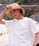 A woman wearing a white t-shirt and the Crochet Raffia Sun Hat tilts its brim with one hand, standing outdoors by a rustic fence against a softly blurred natural background.