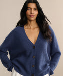 A woman with long dark hair wears the Cropped Cashmere Cocoon Cardigan in deep blue with a V-neck and ribbed texture, standing against a neutral beige background.