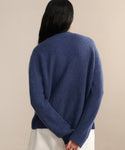 A person with long dark hair, wearing the Cropped Cashmere Cocoon Cardigan in blue and a white skirt, stands facing away with hands clasped behind their back against a neutral background.
