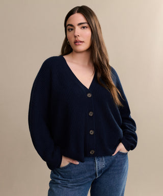 A woman with long brown hair wears the Cropped Cashmere Cocoon Cardigan in navy and blue jeans, standing with her hands in her pockets against a plain beige background.