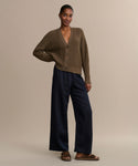 A woman stands against a beige background wearing the Cropped Cotton Cocoon Cardigan in brown, loose navy pants, and brown sandals. One hand is in her pocket, and she looks relaxed.