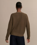 A person with a low bun faces away from the camera, wearing the Cropped Cotton Cocoon Cardigan in olive and dark pants, set against a plain beige background.
