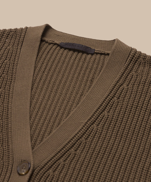 Close-up of the Cropped Cotton Cocoon Cardigan, featuring a brown ribbed knit, V-neckline, buttons, and an inside-collar label, set against a beige background.