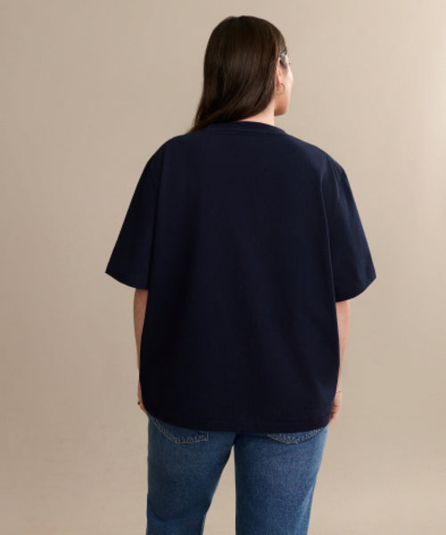 Wearing the relaxed fit Crosby Tee in navy blue premium jersey cotton and blue jeans, a person with long brown hair stands facing away from the camera against a plain beige background.