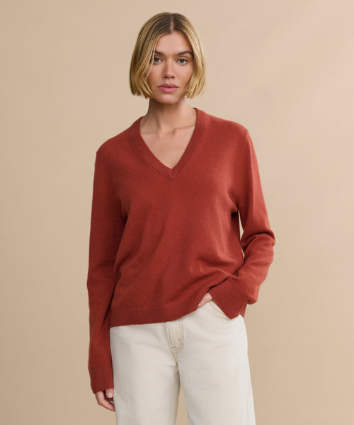 A woman with a bob haircut wears the rust-colored, relaxed fit Flynn Cashmere Sweater and cream pants, standing against a beige background and looking directly at the camera.