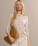 A woman with long blonde hair wears the Grayson Pullover and light pants, holding a large mustard clutch, standing against a plain beige background.
